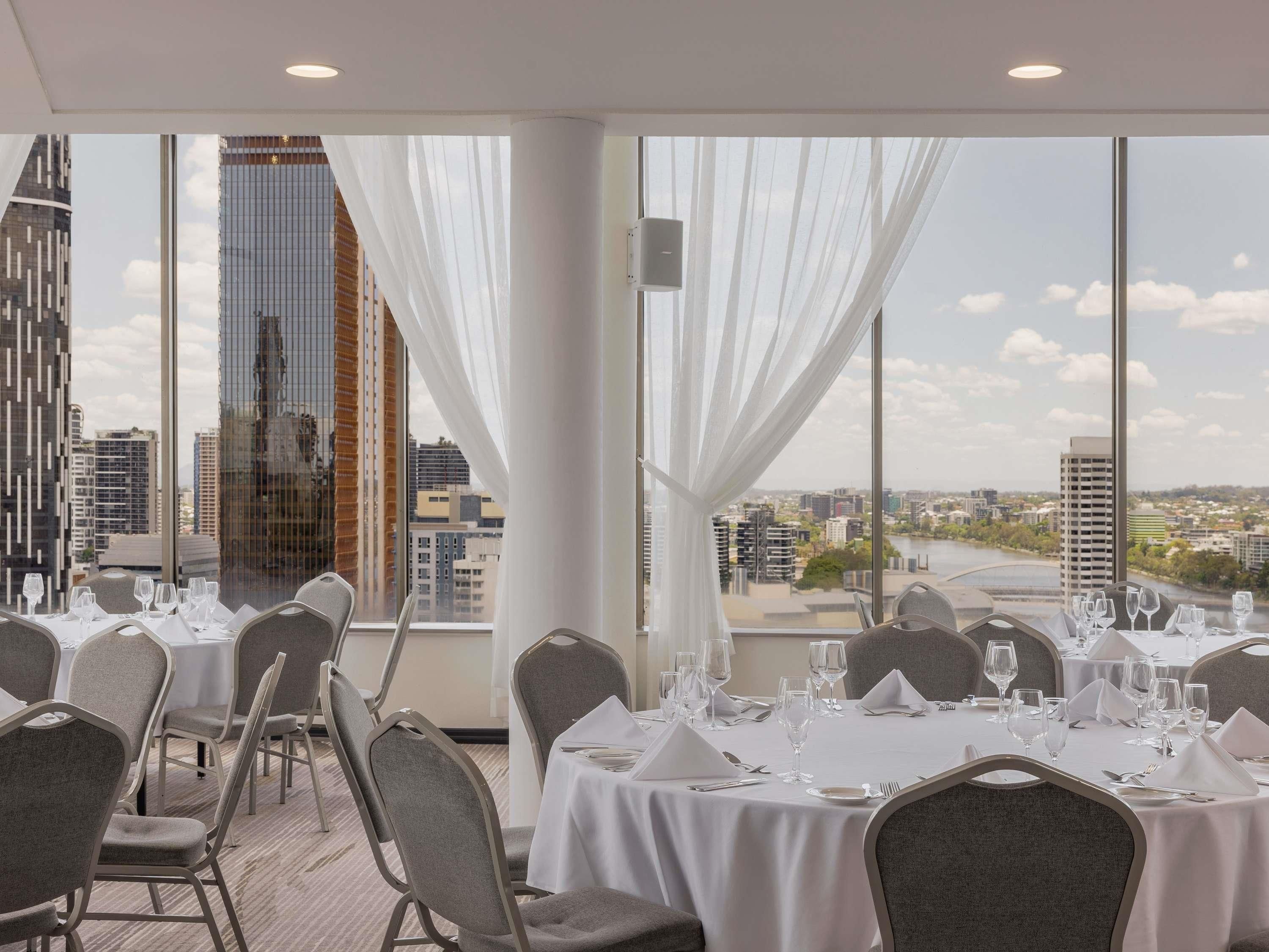 Un comedor con mesas dispuestas para el servicio, ventanas grandes con cortinas y vistas a la ciudad.