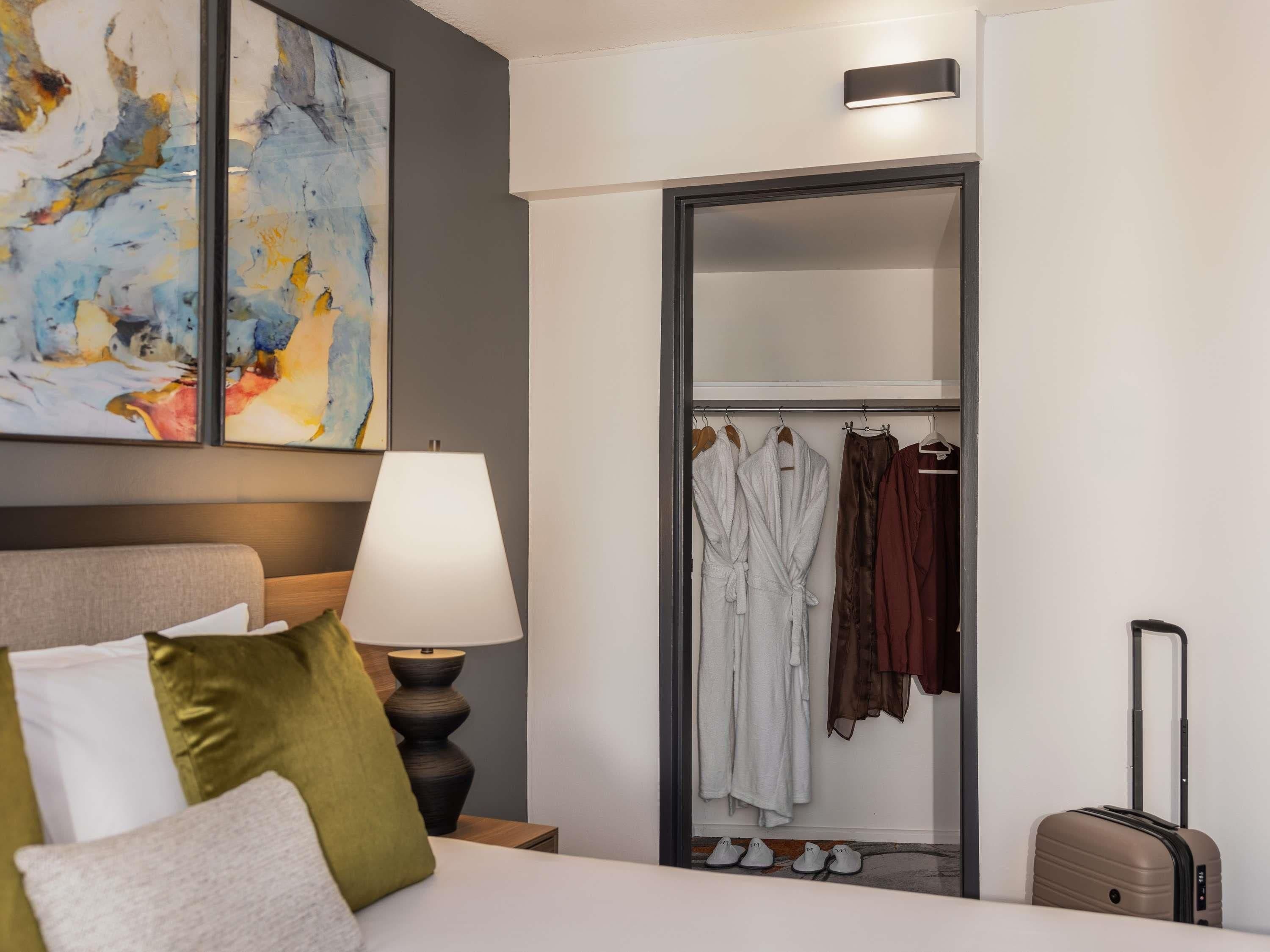 Une chambre d’hôtel avec un lit, une lampe de chevet, une armoire avec des vêtements suspendus et une valise.