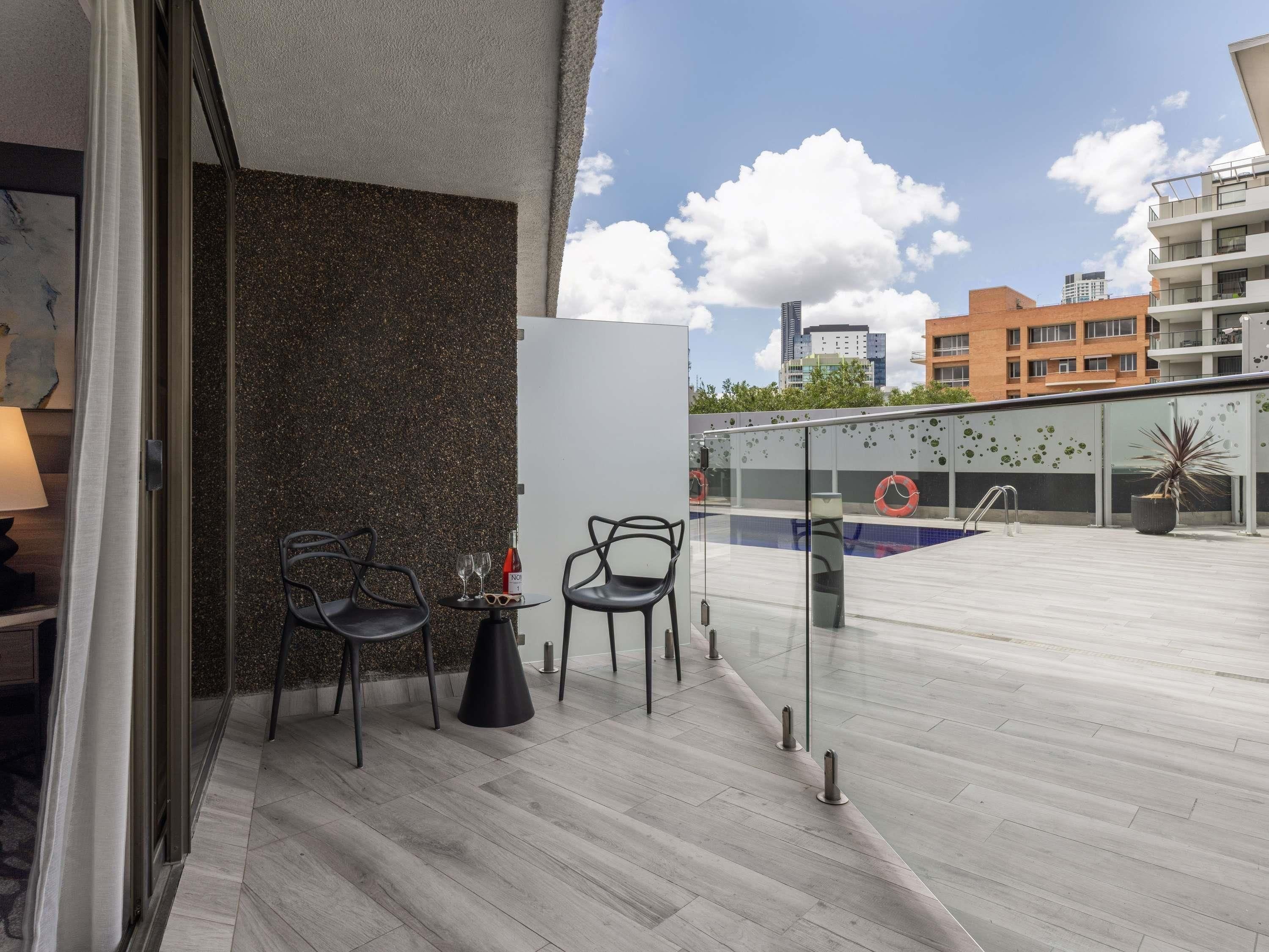 Un espacio exterior moderno con dos sillas y una mesa pequeña, barandilla de cristal con vistas a una piscina y un paisaje urbano de fondo.