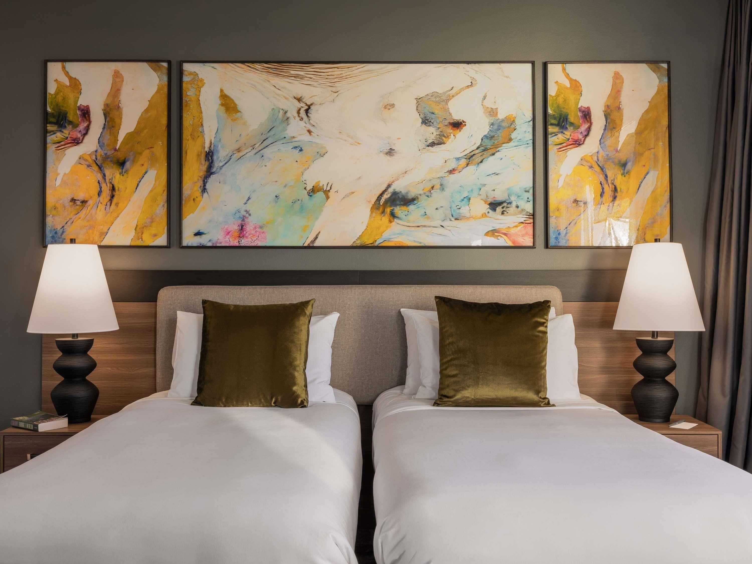 A hotel room with two beds, a large abstract painting above, and two bedside lamps.