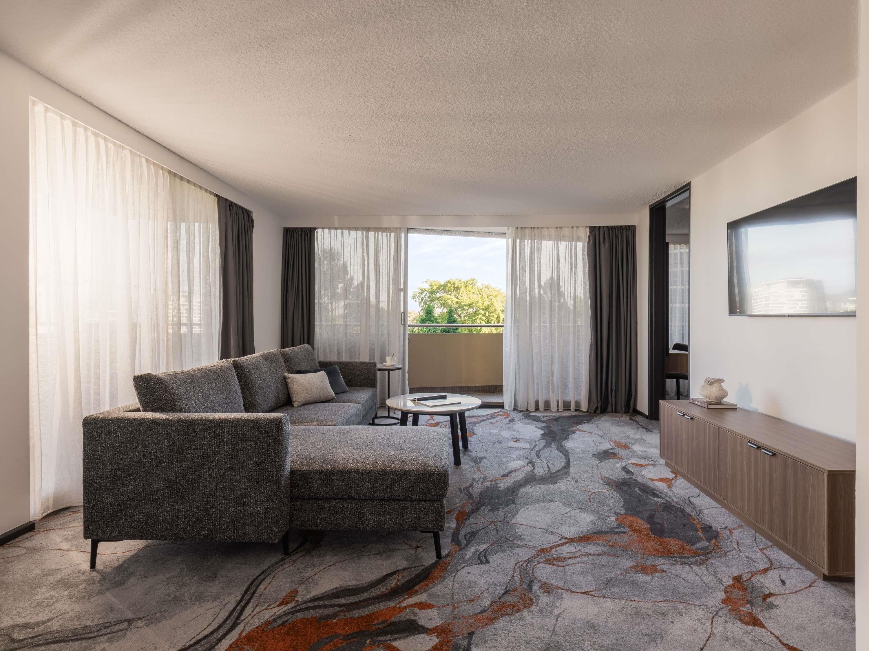 A modern hotel room with a grey sofa, a small round table, and a view of the outdoors through a sliding door.