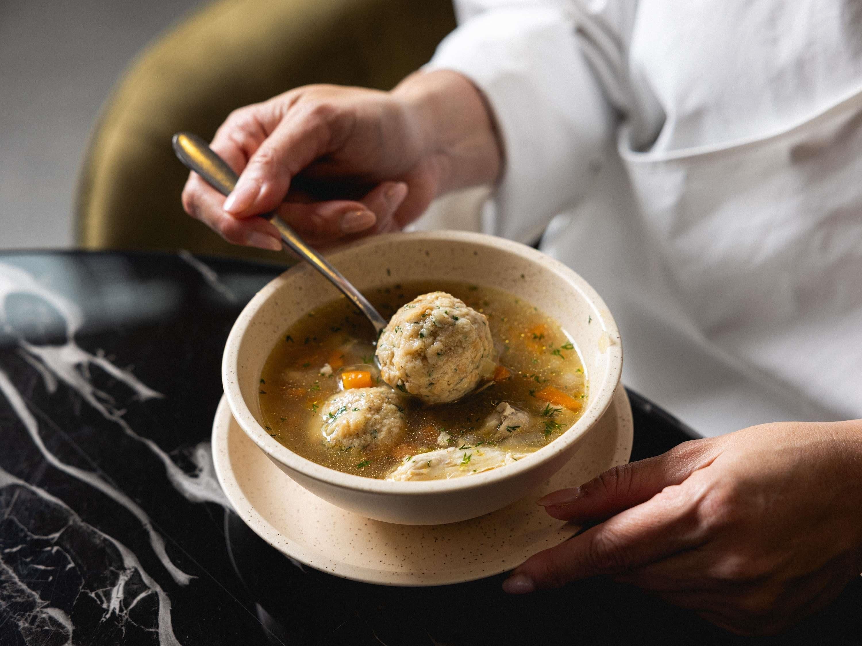 Una persona sosteniendo un tazón de sopa con albóndigas, usando una cuchara para comer.