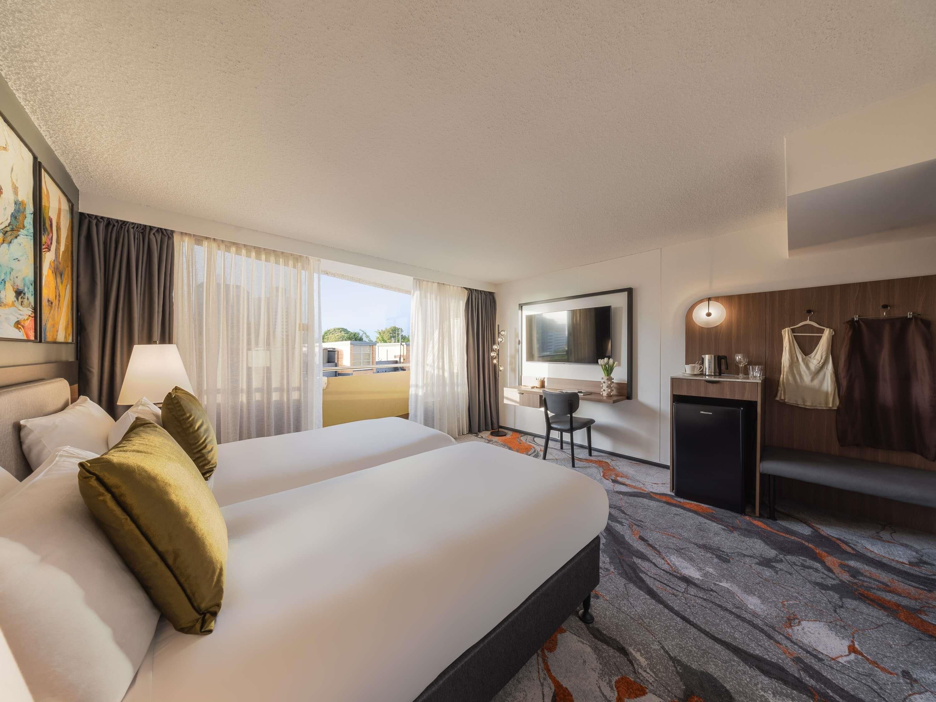 A hotel room with a bed, a desk, a mirror, a TV, and a view of the outside.