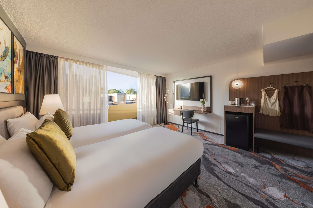 A hotel room with a bed, a desk, a mirror, a TV, and a view of the outside.