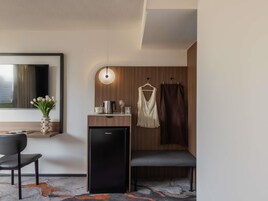 Een moderne hotelkamer met een bureau, stoel, kleine zwarte koelkast, hanglamp, spiegel en raam met uitzicht.