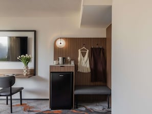Habitación de hotel moderna con escritorio, silla, refrigerador negro pequeño, lámpara colgante, espejo y ventana con vistas.