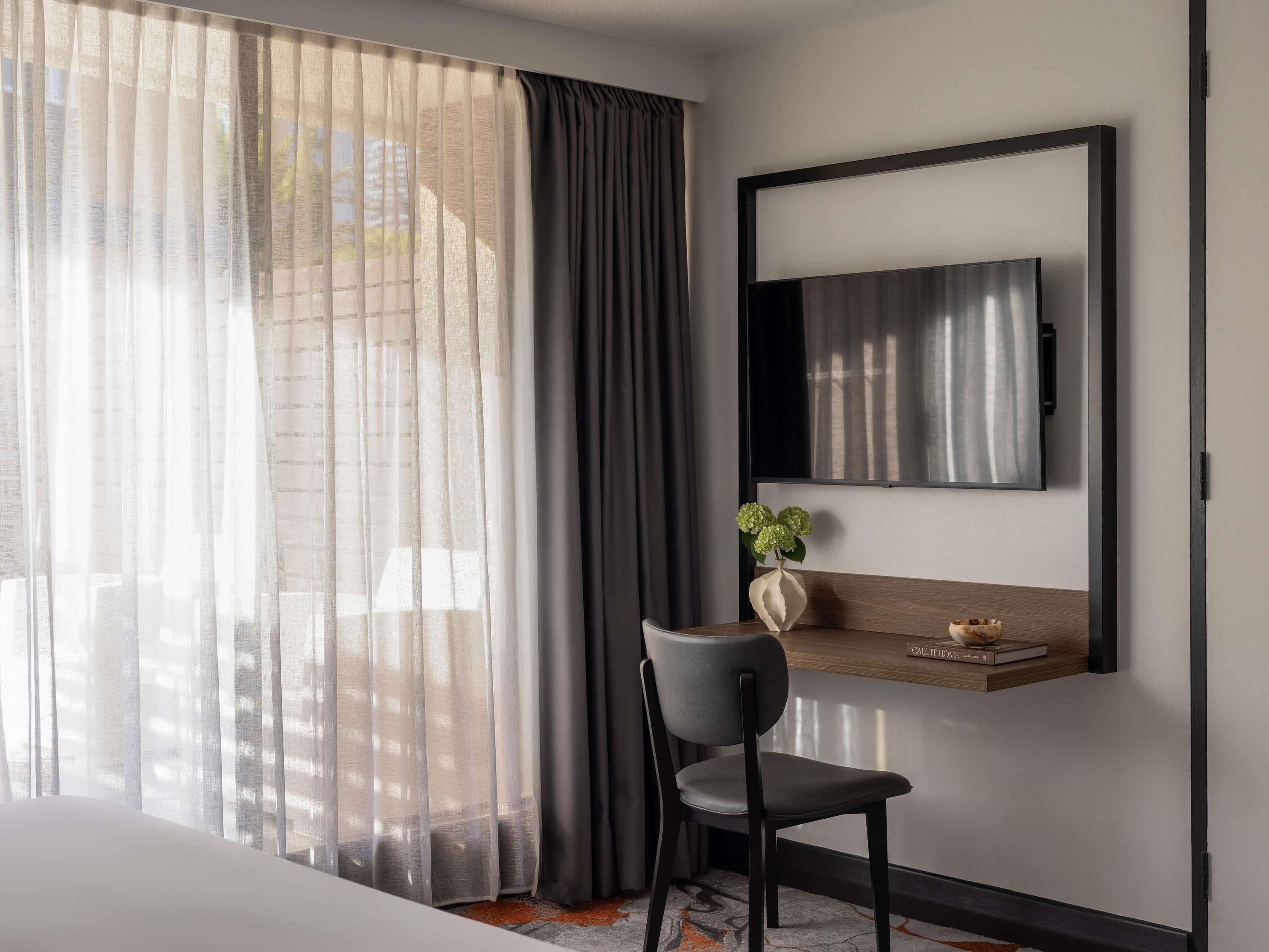 A hotel room with a large window, a chair, a small table with a vase, and a mounted TV.