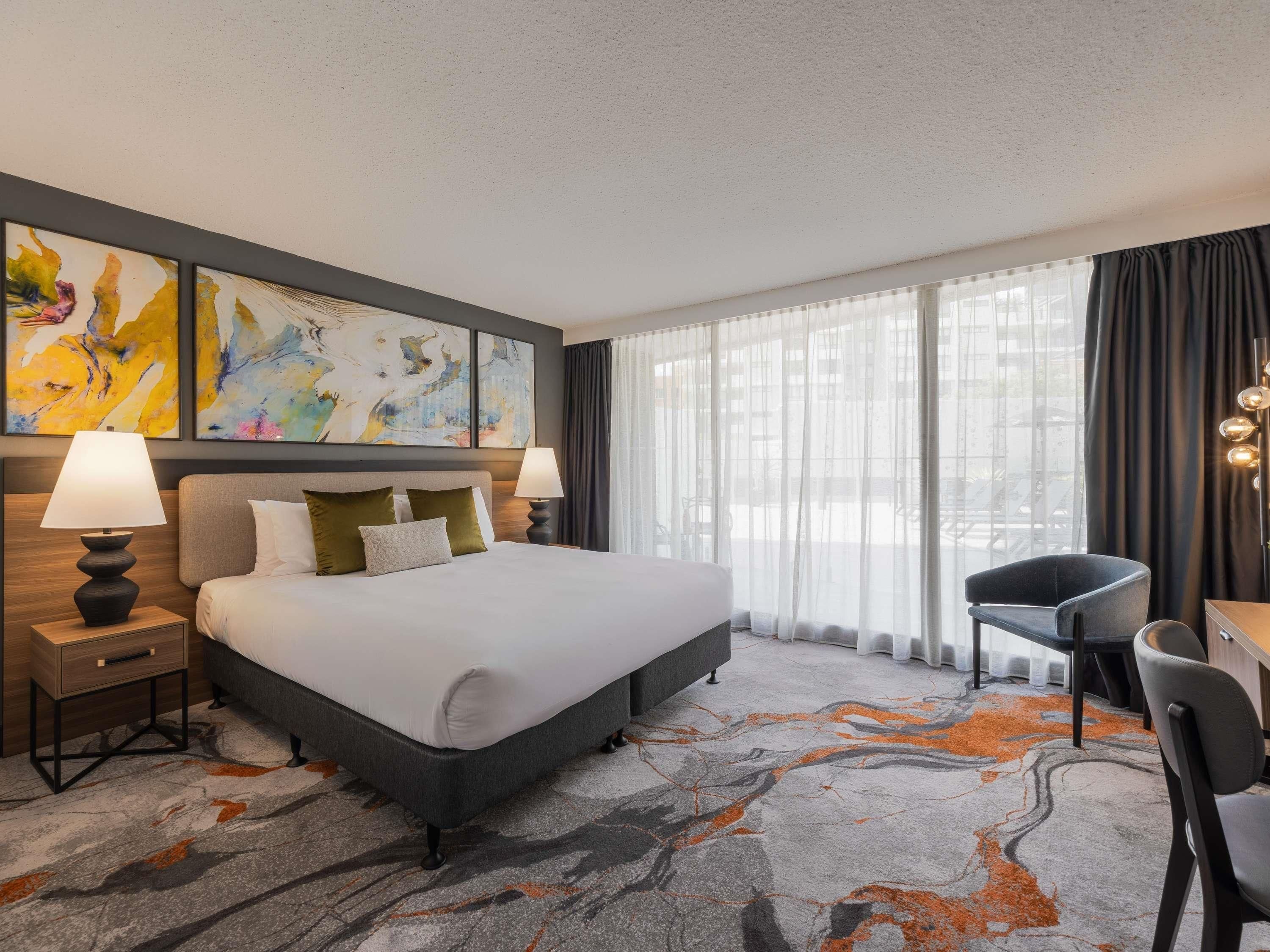 A hotel room with a large bed, two bedside tables with lamps, a desk with a chair, and a large window with sheer curtains.