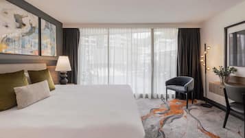 Ett hotellrum med en säng, ett skrivbord, en stol, en lampa, en tavla och utsikt genom fönstret.