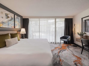 A hotel room with a bed, a desk, a chair, a lamp, a painting, and a view through the window.