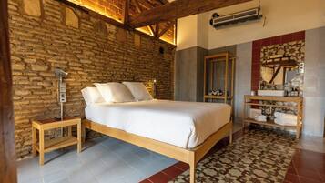 Un dormitorio con pared de piedra, vigas de madera, cama con ropa blanca, mesita de noche, espejo y estante con toallas.