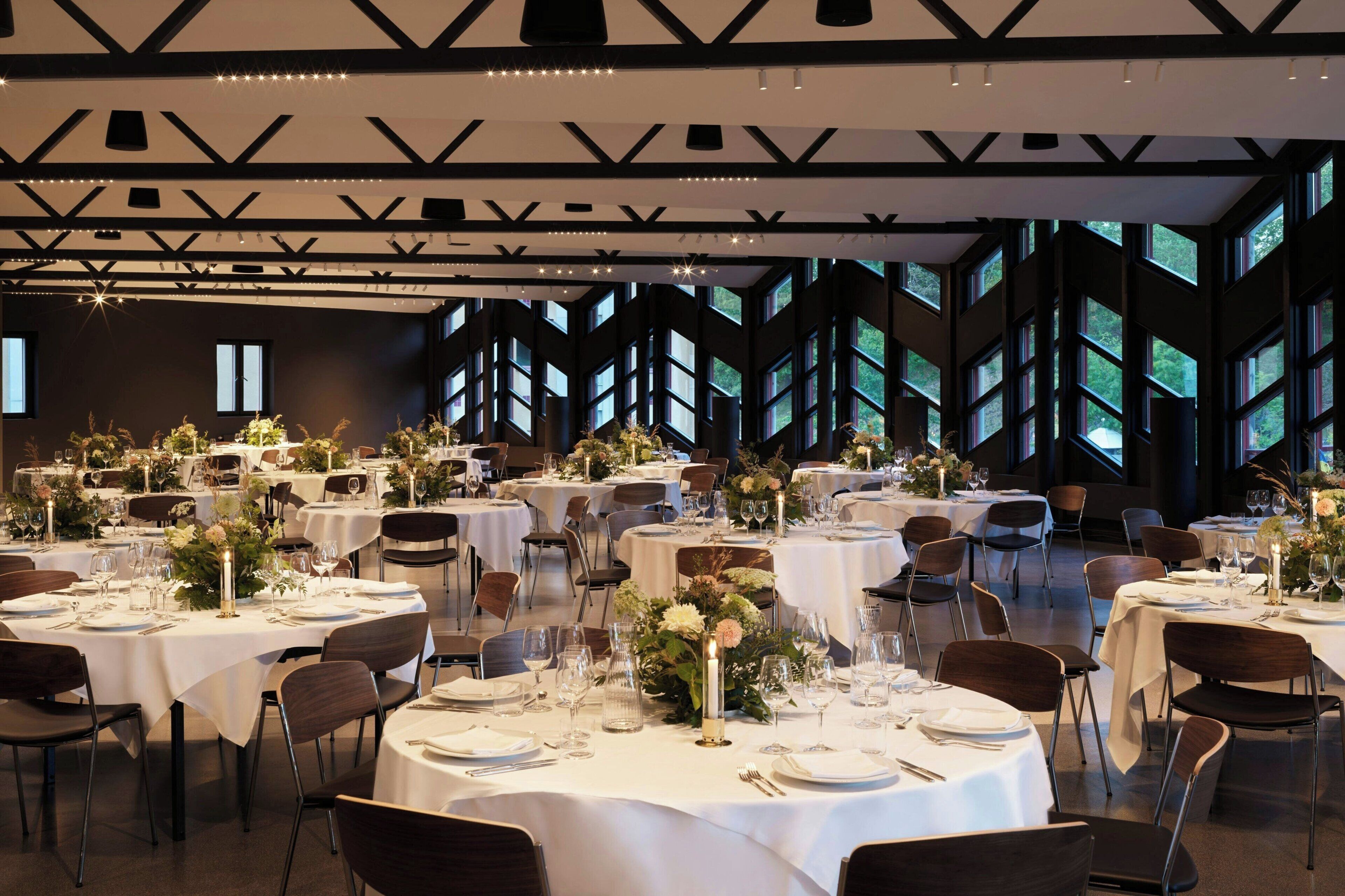 A spacious banquet hall with multiple round tables set for dining, featuring white tablecloths and floral centerpieces.