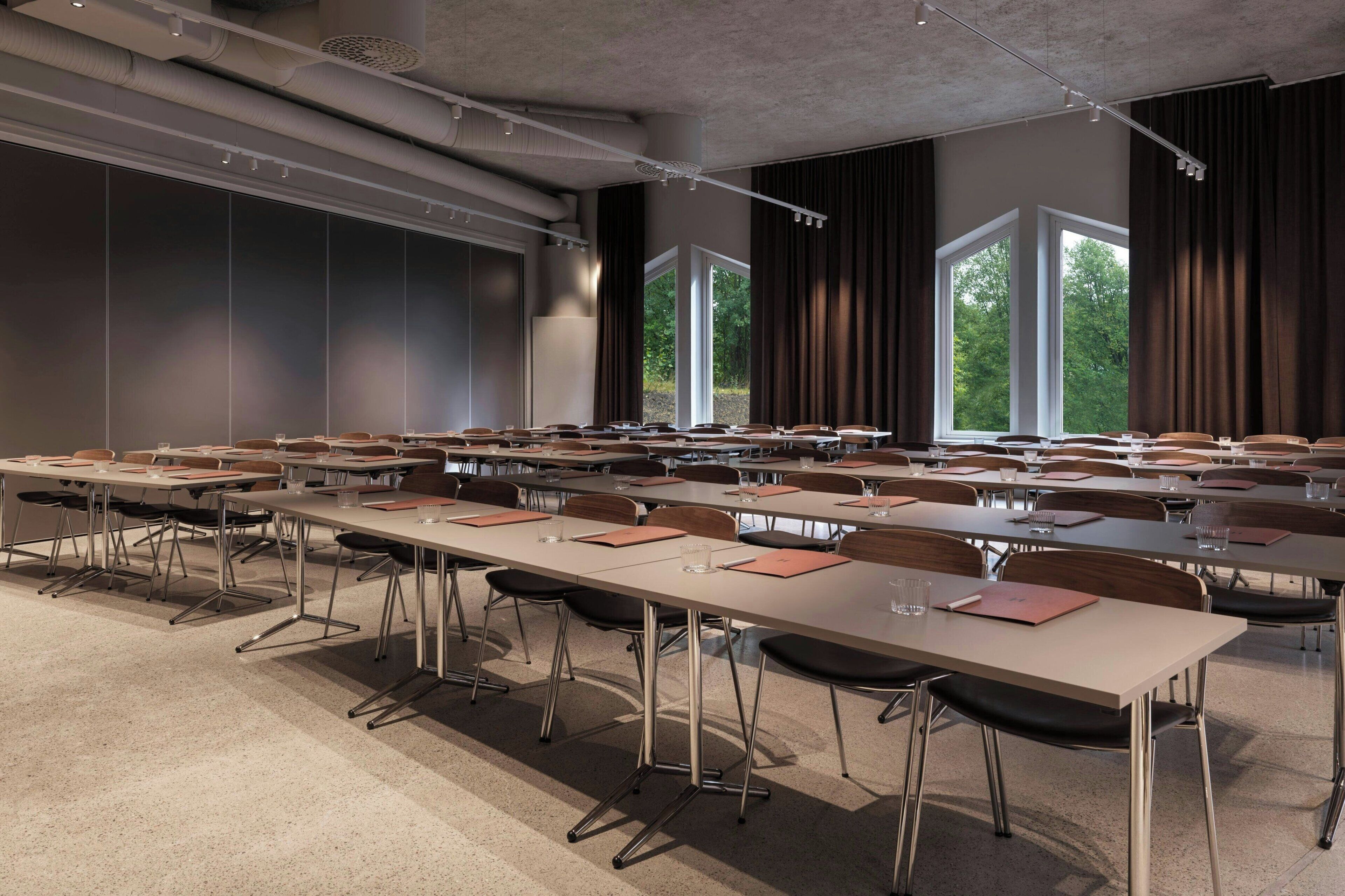 A conference room with multiple tables and chairs, large windows with curtains, and a high ceiling with visible ductwork.