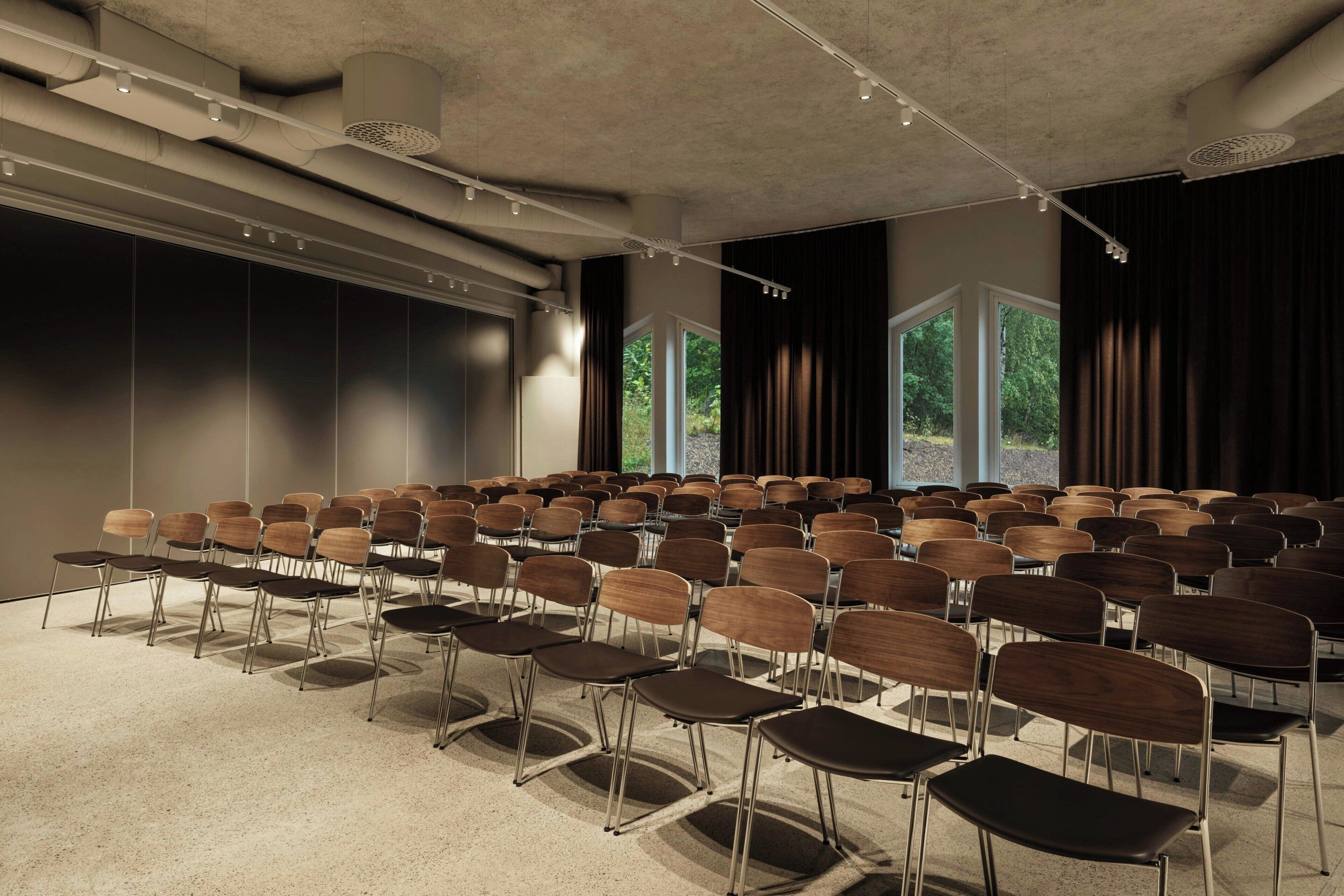 Konferenssihuone, jossa on riveissä tuoleja, suuret ikkunat ja katto, jossa ilmanvaihtoputket ovat näkyvillä.
