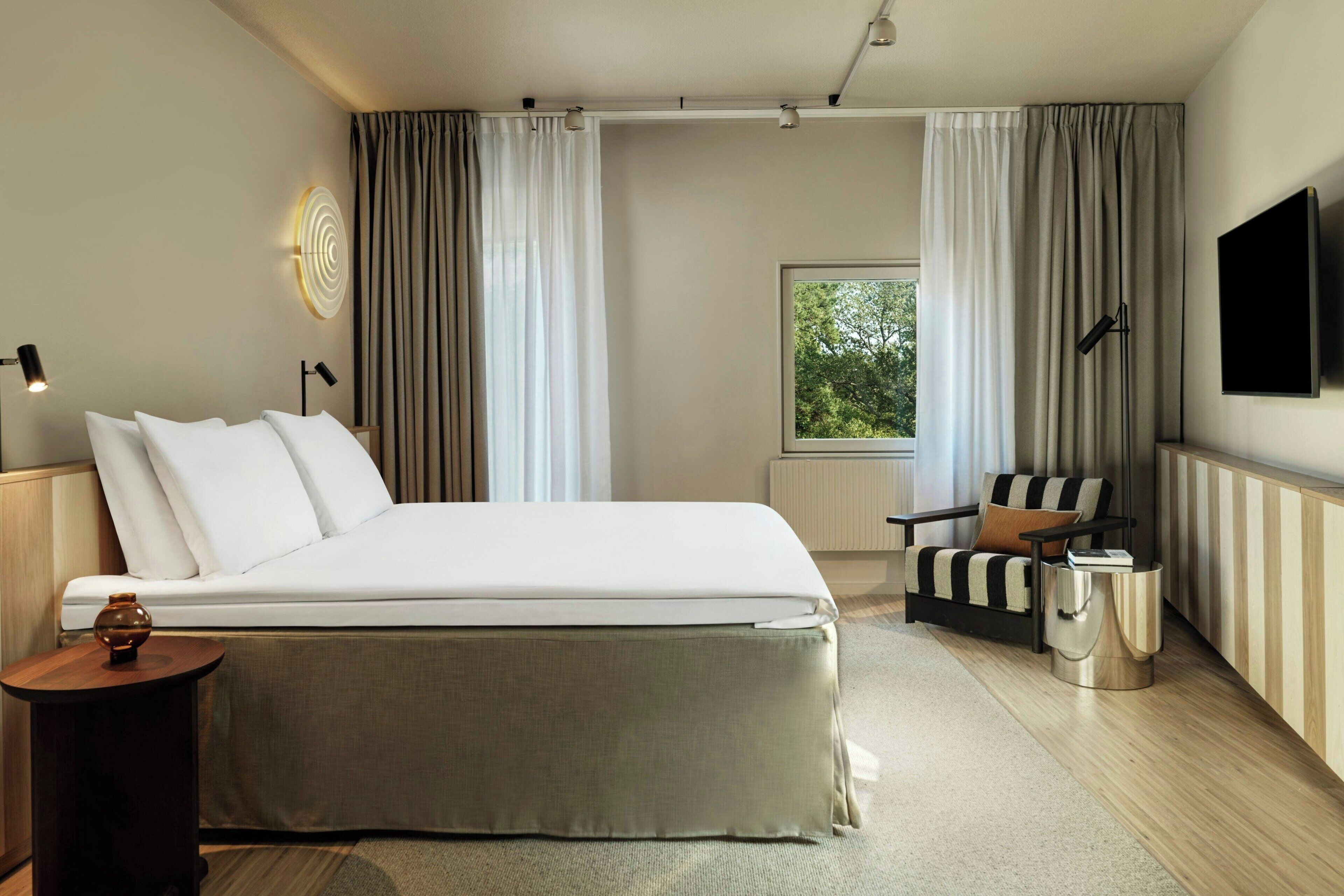 Quarto de hotel moderno com uma cama grande, área de estar com uma poltrona e uma mesa pequena, televisão de tela plana na parede e uma janela com cortinas.