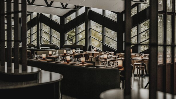 A modern restaurant with high ceilings, large windows, and a dark interior. Tables are set with candles and wine glasses. - Hagastrand, Autograph Collection (Solna)