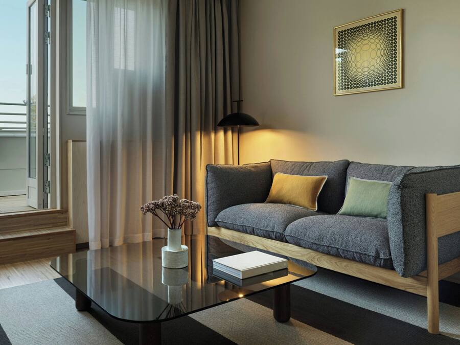A modern living room with a sofa, a glass coffee table, and a view of the outdoors through a window with sheer curtains.