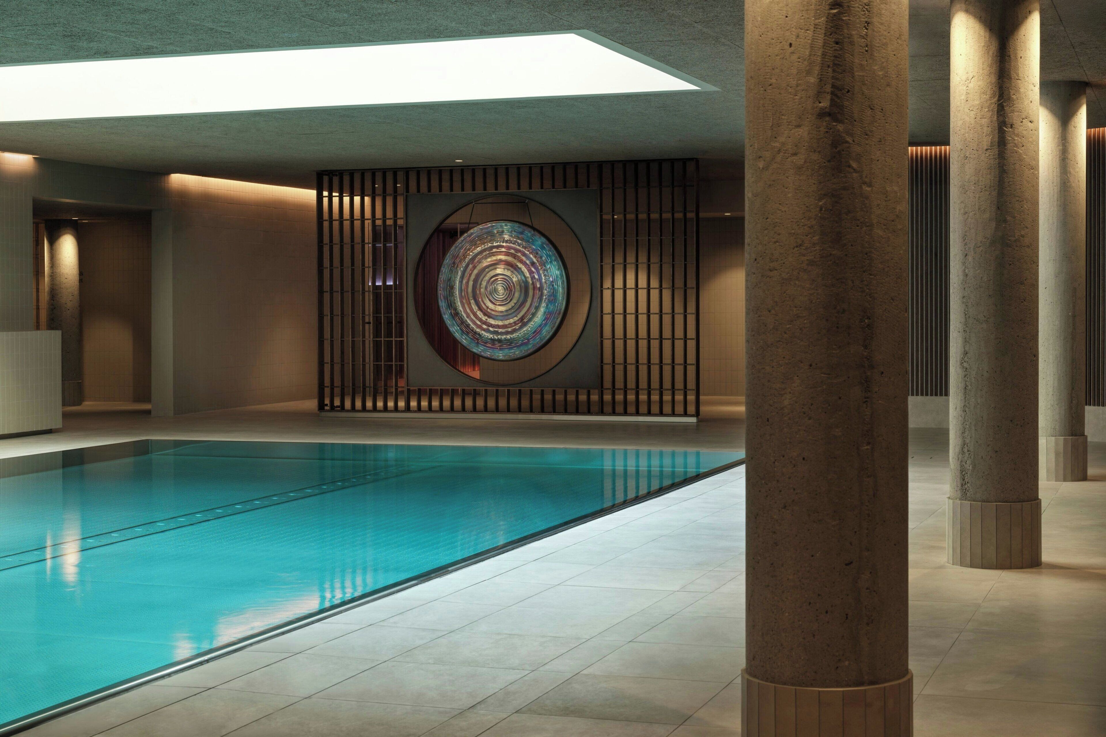 A modern indoor swimming pool with a geometric wall art piece, concrete pillars, and a skylight.