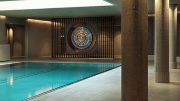 A modern indoor swimming pool with a geometric wall art piece, concrete pillars, and a skylight.