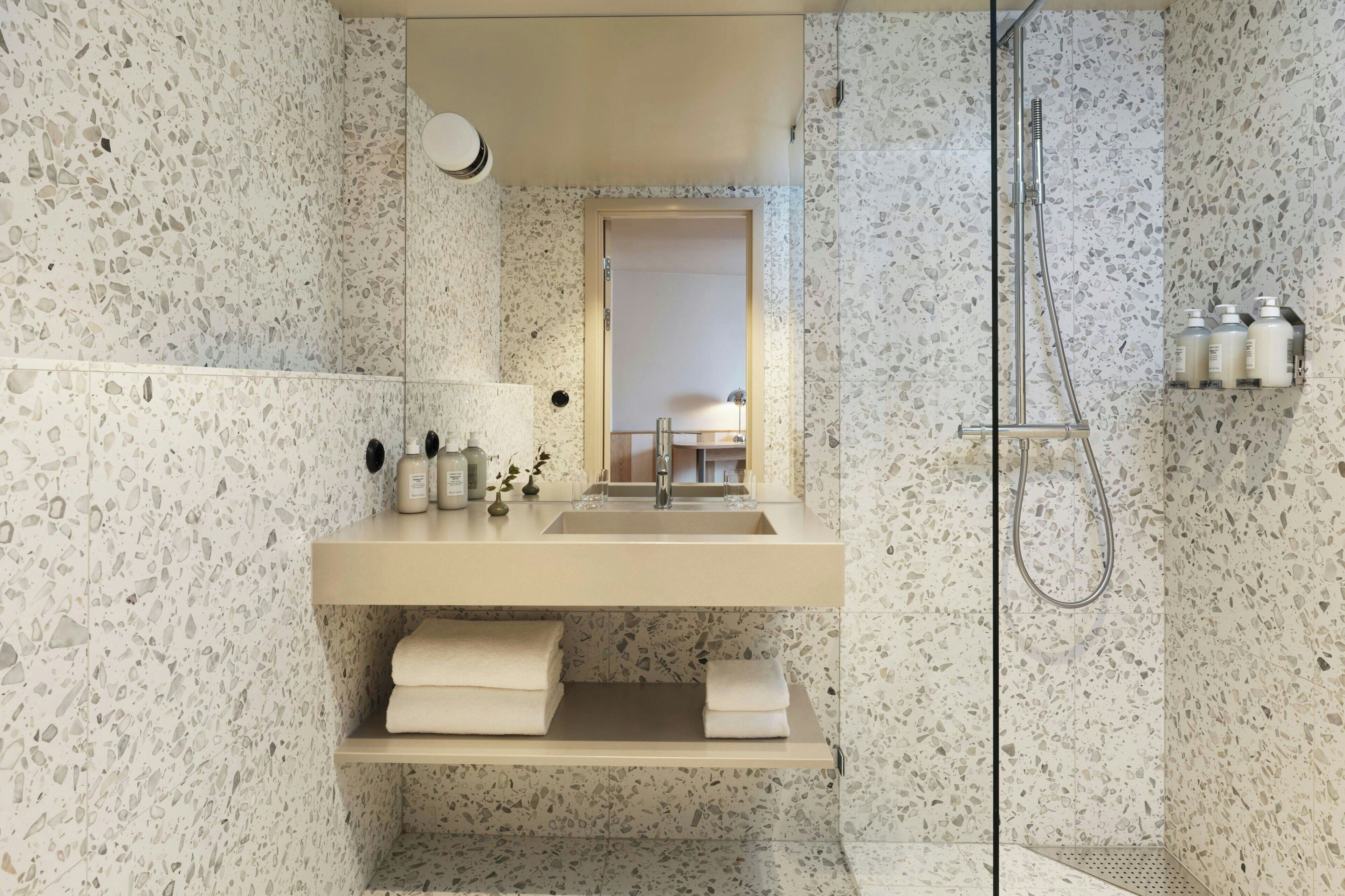 A bathroom with a terrazzo floor, a sink with a mirror, a shower, and a shelf with toiletries.