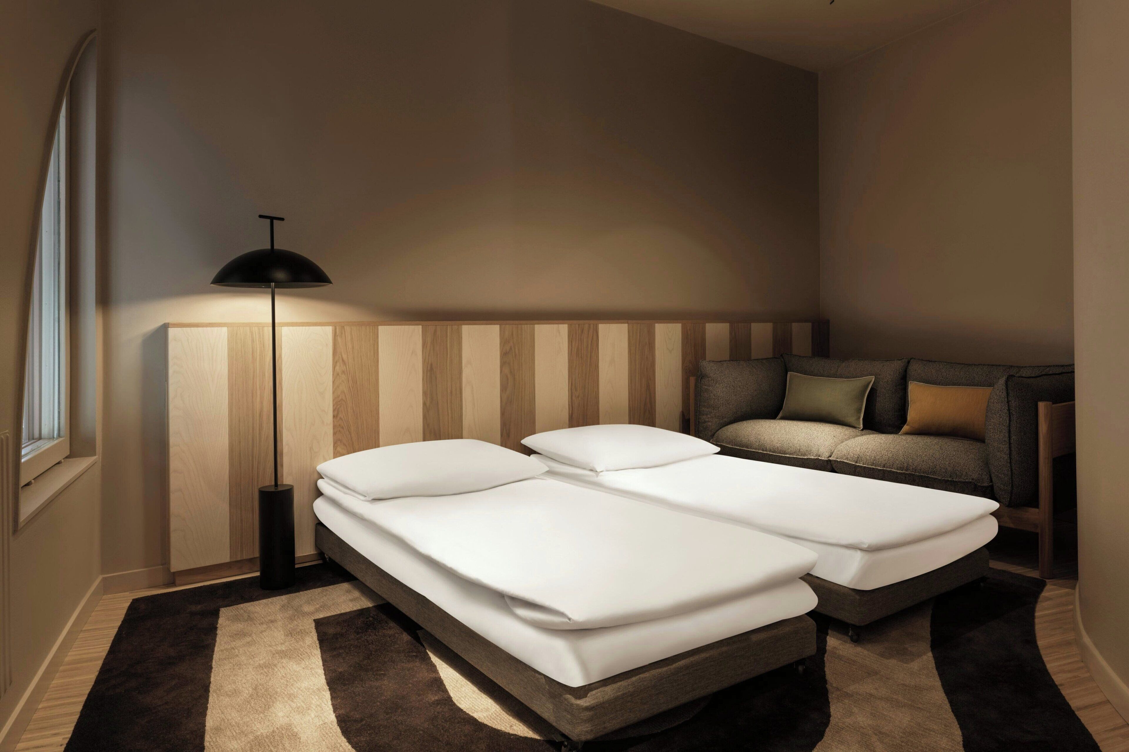 Une chambre moderne avec deux lits, une lampe de chevet, un canapé et une tête de lit en bois.