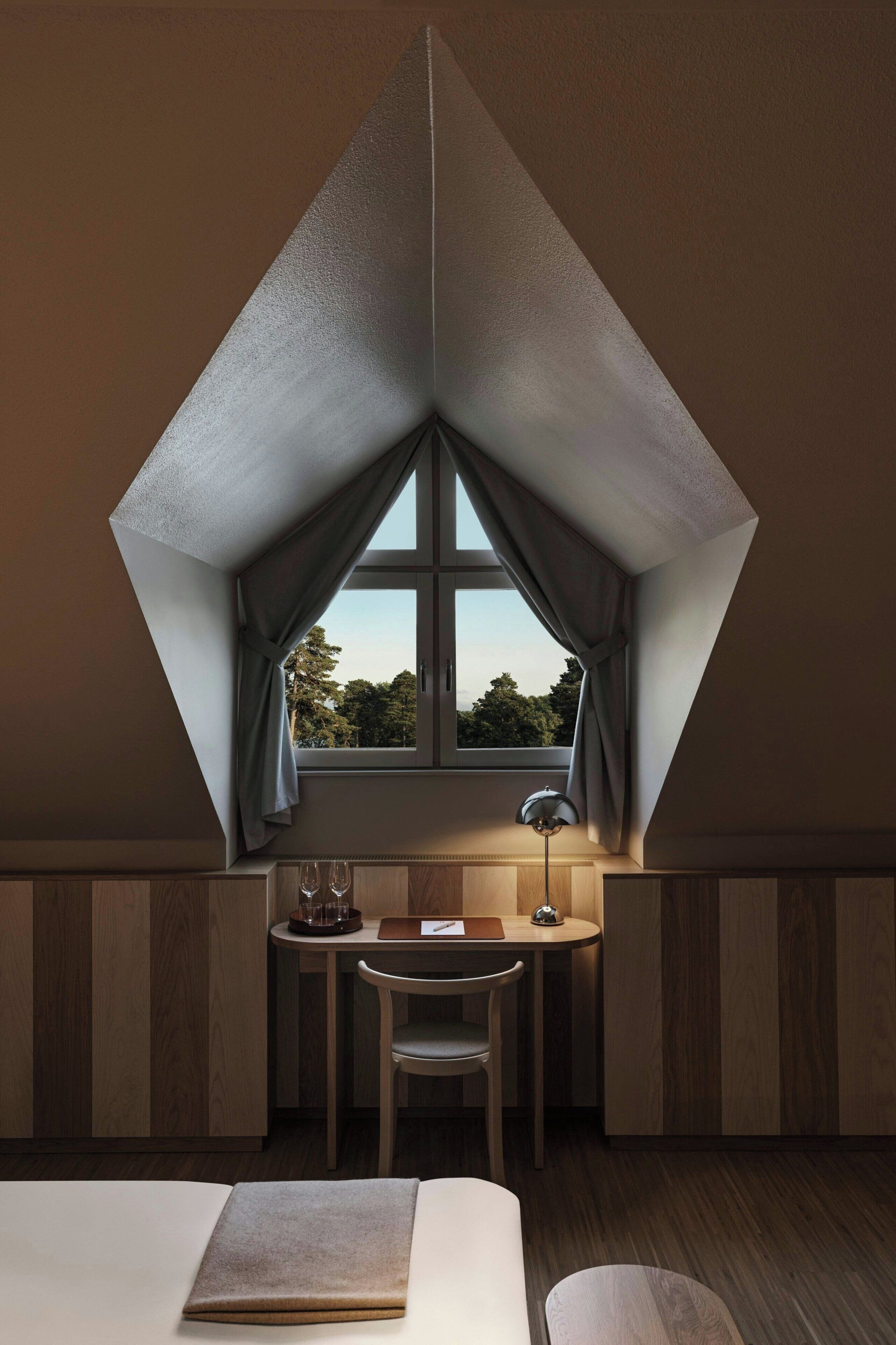 A room with a triangular ceiling, a small table with a chair, a lamp, and a bed.