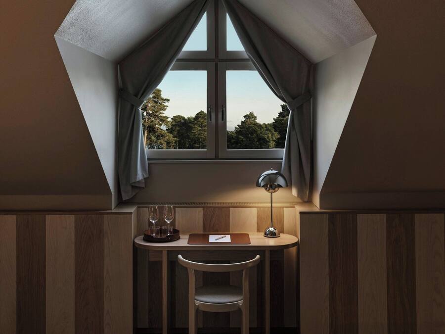 A room with a triangular ceiling, a small table with a chair, a lamp, and a bed.