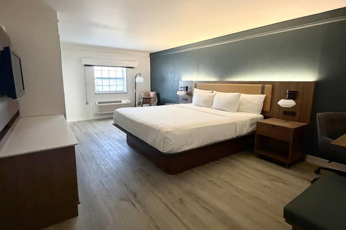 A hotel room with a large bed, bedside tables, a desk, a chair, a window, and an air conditioning unit.