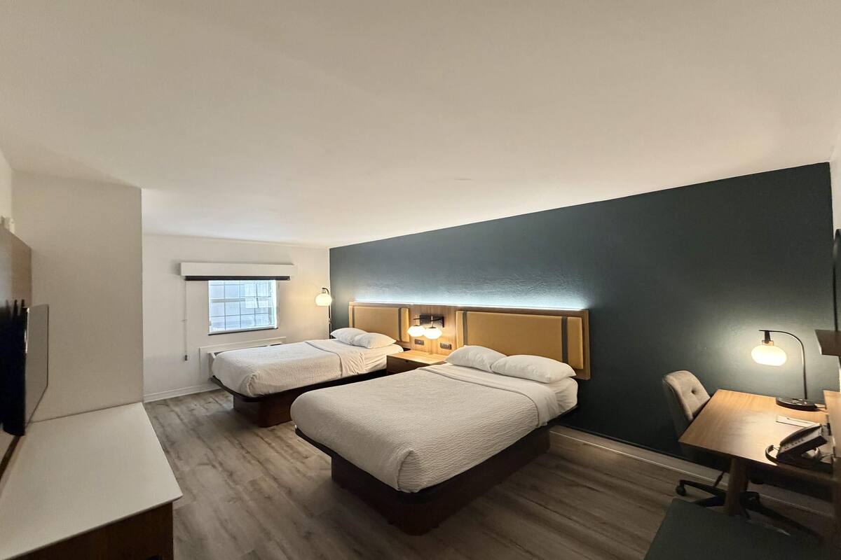 A hotel room with two beds, a desk, a chair, and a small table. There is a window providing natural light and a wall-mounted lamp.