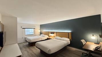 A hotel room with two beds, a desk, a chair, and a small table. There is a window providing natural light and a wall-mounted lamp.