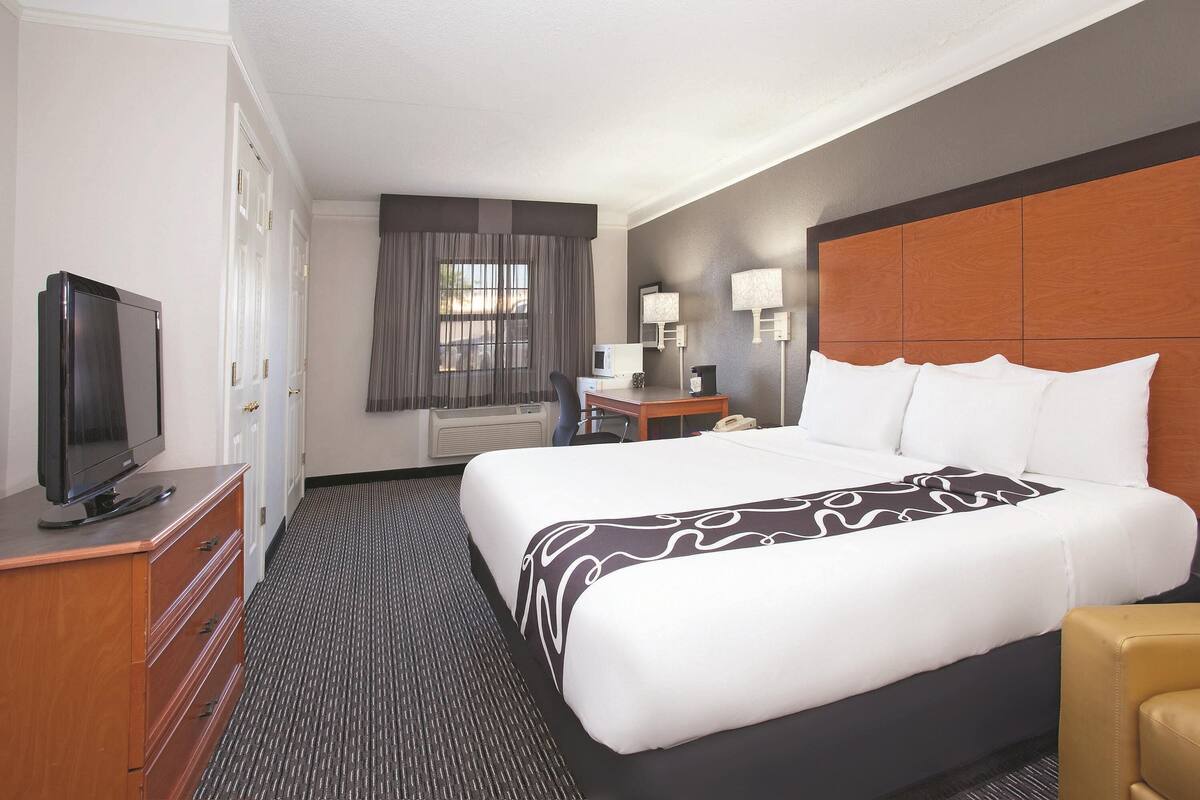 A hotel room with a large bed, a television on a dresser, a desk with a lamp, and a window with curtains.