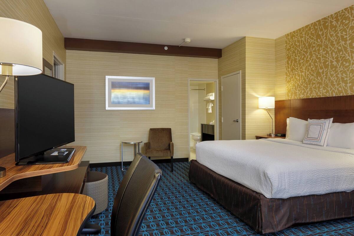 A hotel room with a bed, a desk with a television, a chair, a small table, and a bathroom visible through an open door.