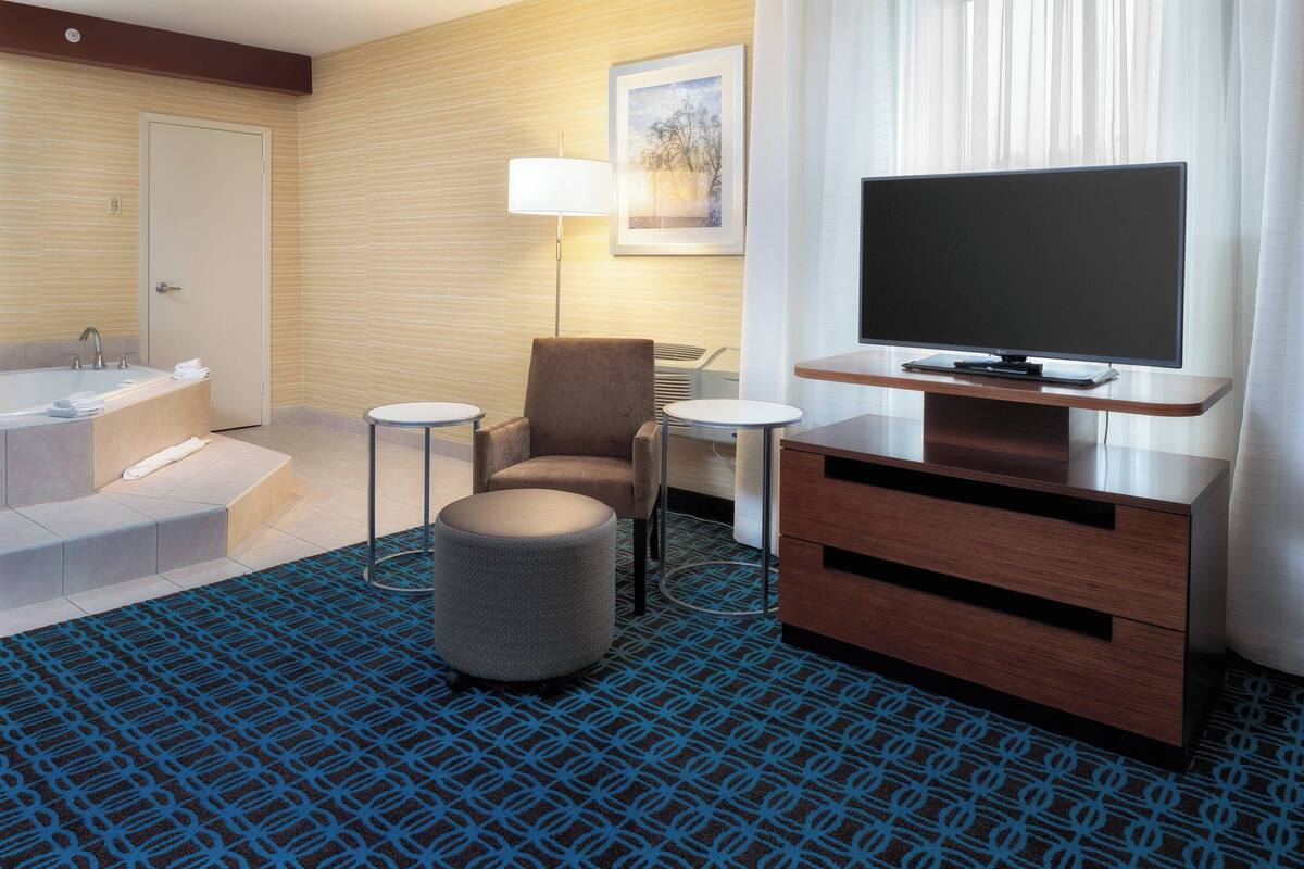 A hotel room with a flat-screen TV, a sofa, a small round ottoman, a side table, a lamp, a bathroom with a bathtub, and a patterned carpet.