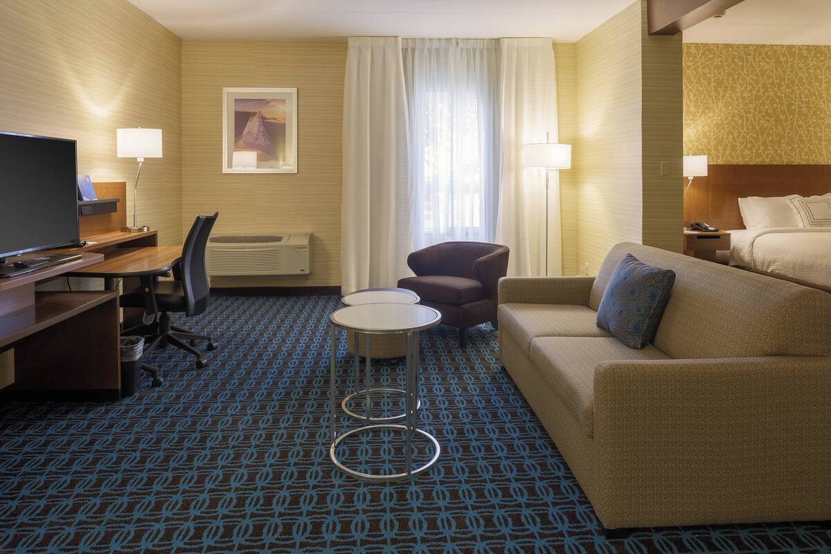 A hotel room with a bed, a desk with a computer, a chair, a small table, and a TV.