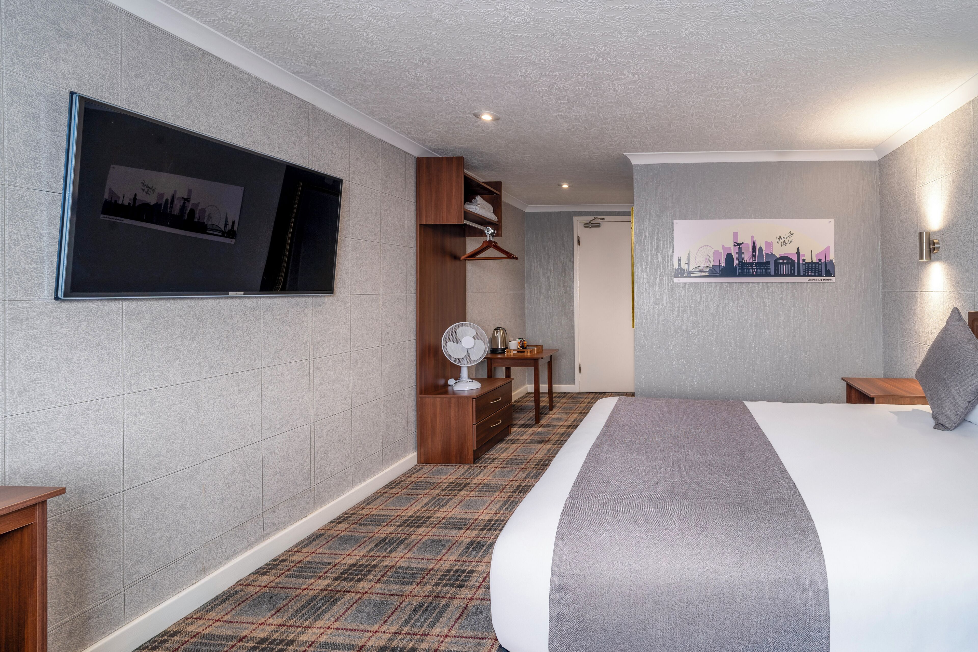 Habitación de hotel con una cama grande, un televisor de pantalla plana en la pared, un escritorio con una silla, un armario y un cuadro de paisaje urbano.