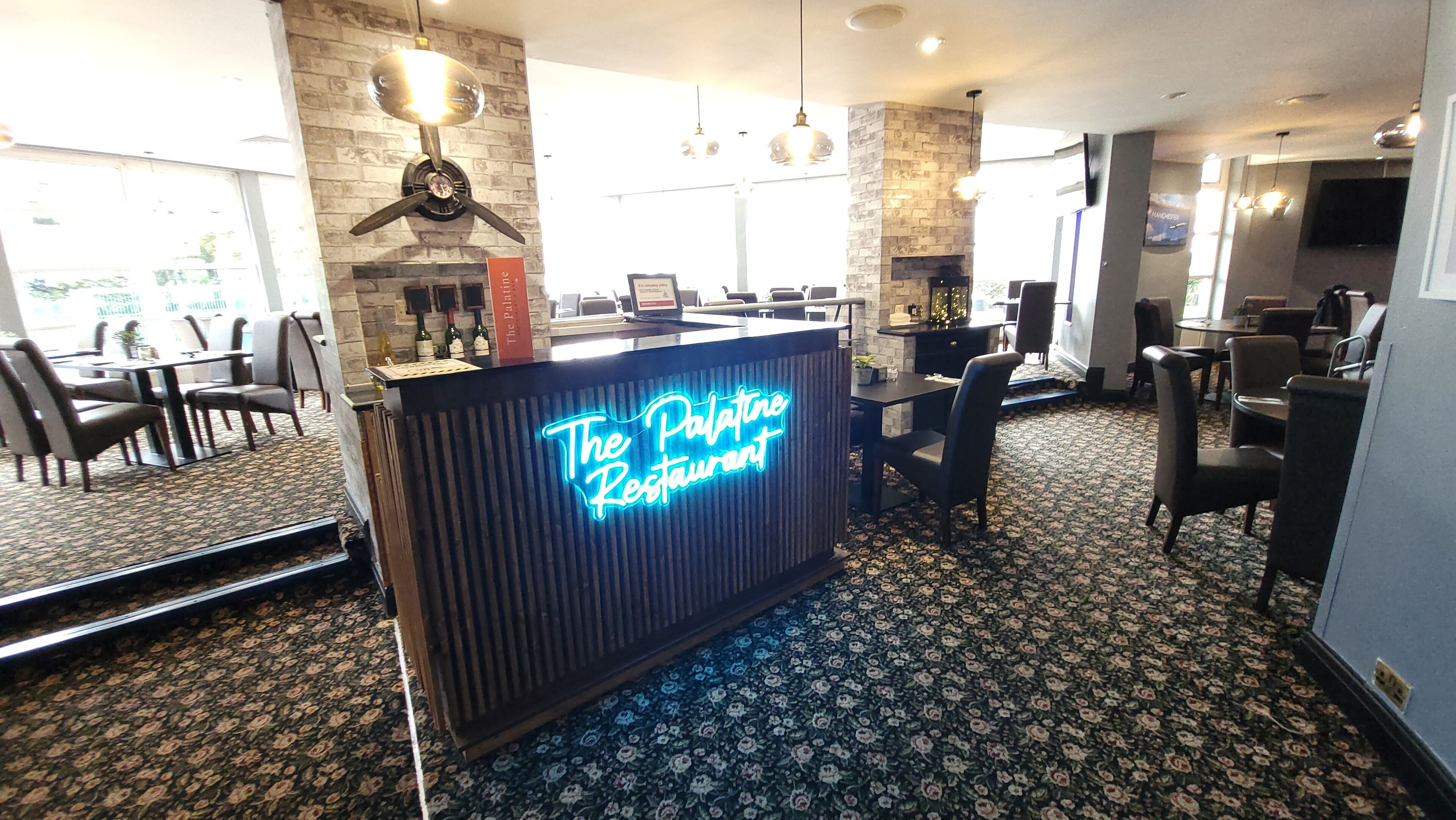 L’intérieur d’un restaurant, avec un panneau lumineux au néon, un comptoir d’accueil en bois, un tapis à motifs et un espace repas comprenant des chaises et des tables.
