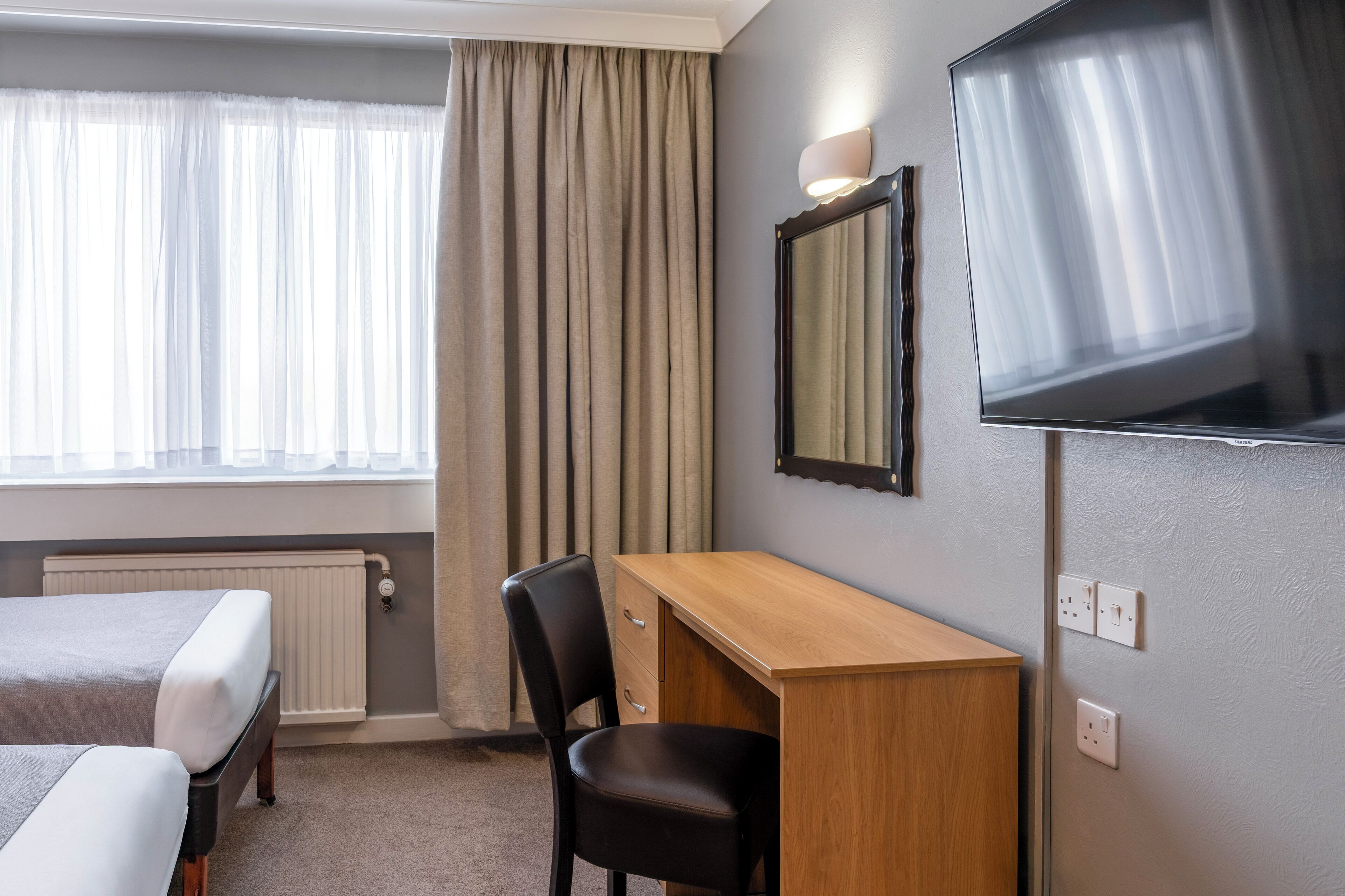 A hotel room with two beds, a desk, a chair, a television, a mirror, and a window with curtains.