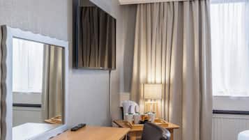 Habitación de hotel con un escritorio de madera, una silla, un televisor en la pared, una lámpara y una ventana con cortinas.