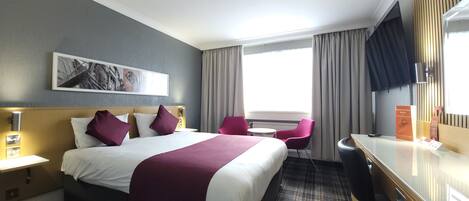 A hotel room with a bed, two chairs, a desk, a window with curtains, a wall-mounted artwork, and a room service menu stand.