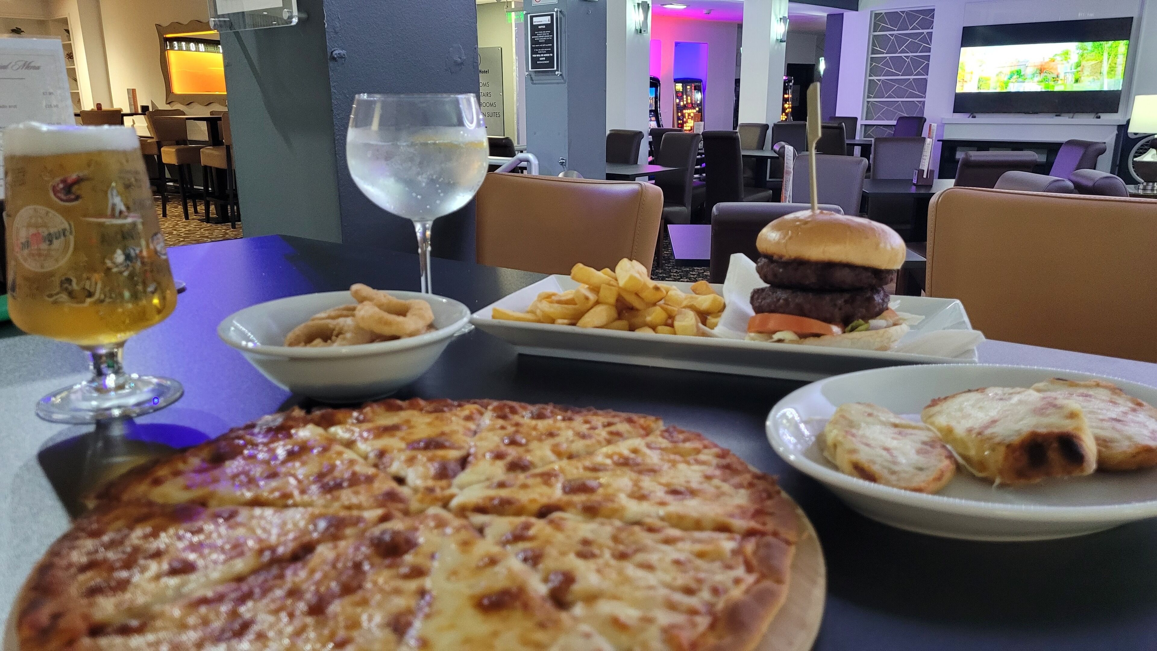 A dining table with a pizza, a burger, fries, onion rings, and a glass of beer.