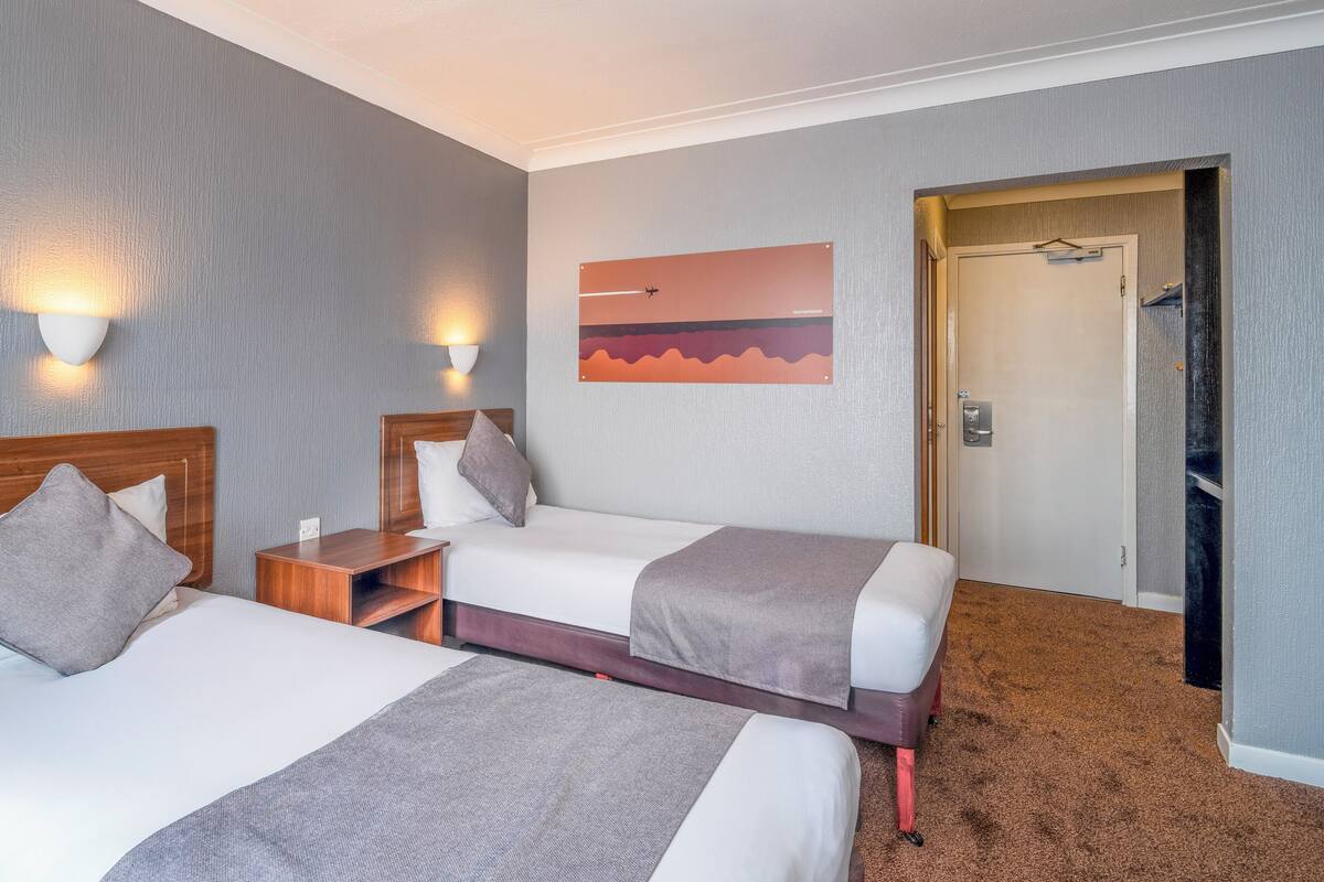 Habitación de hotel con dos camas, una mesita de noche, una lámpara en la pared y una puerta que da a un armario.