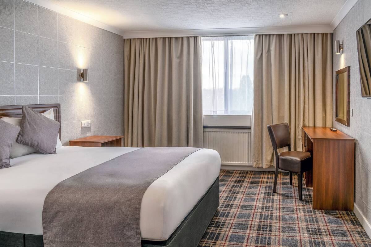 Una habitación de hotel con una cama grande, un escritorio, una silla y un ventanal con cortinas.