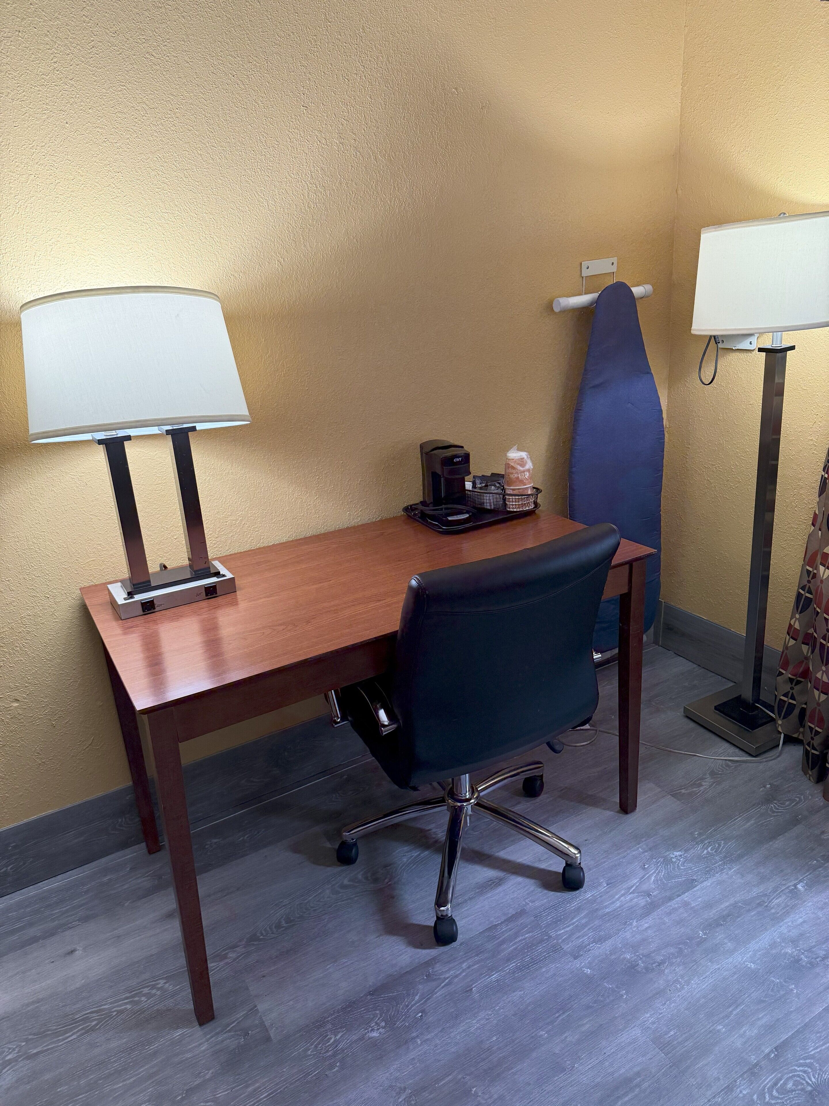 A hotel room with a wooden desk, a black office chair, two wall-mounted lamps, a coffee maker, and a blue cushioned chair.