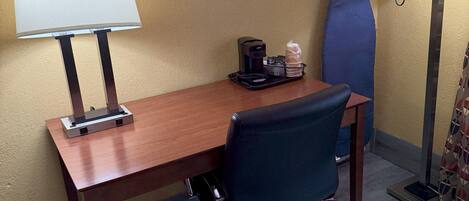 A hotel room with a wooden desk, a black office chair, two wall-mounted lamps, a coffee maker, and a blue cushioned chair.