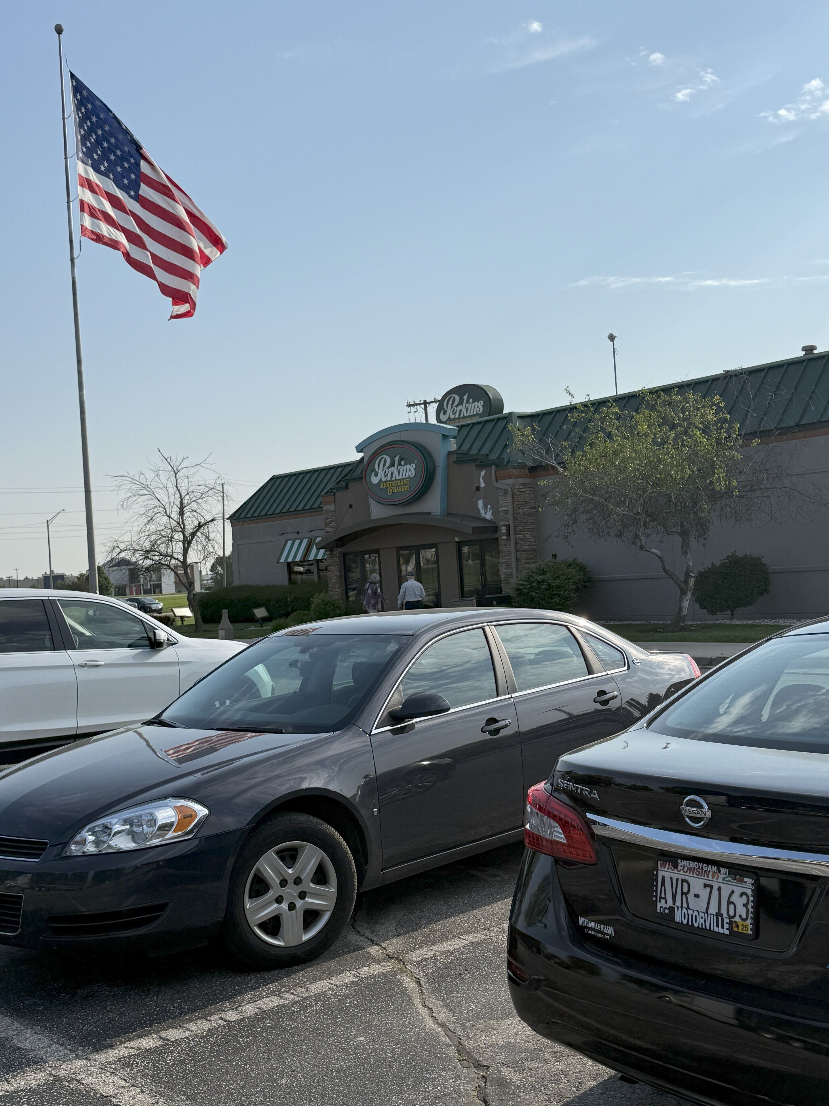 Un estacionamiento con autos, el edificio de un restaurante Perkins y una bandera estadounidense.