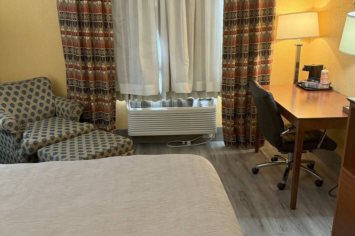 A hotel room with a bed, a desk, a chair, an air conditioner, and curtains.
