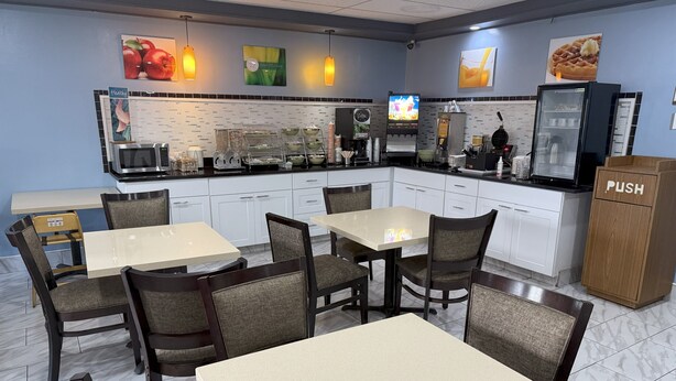 Un restaurante con un interior moderno y luminoso, que presenta una zona de comedor central con mesas y sillas, una cocina con electrodomésticos y un refrigerador, y decoraciones en las paredes con imágenes de frutas y gofres.
