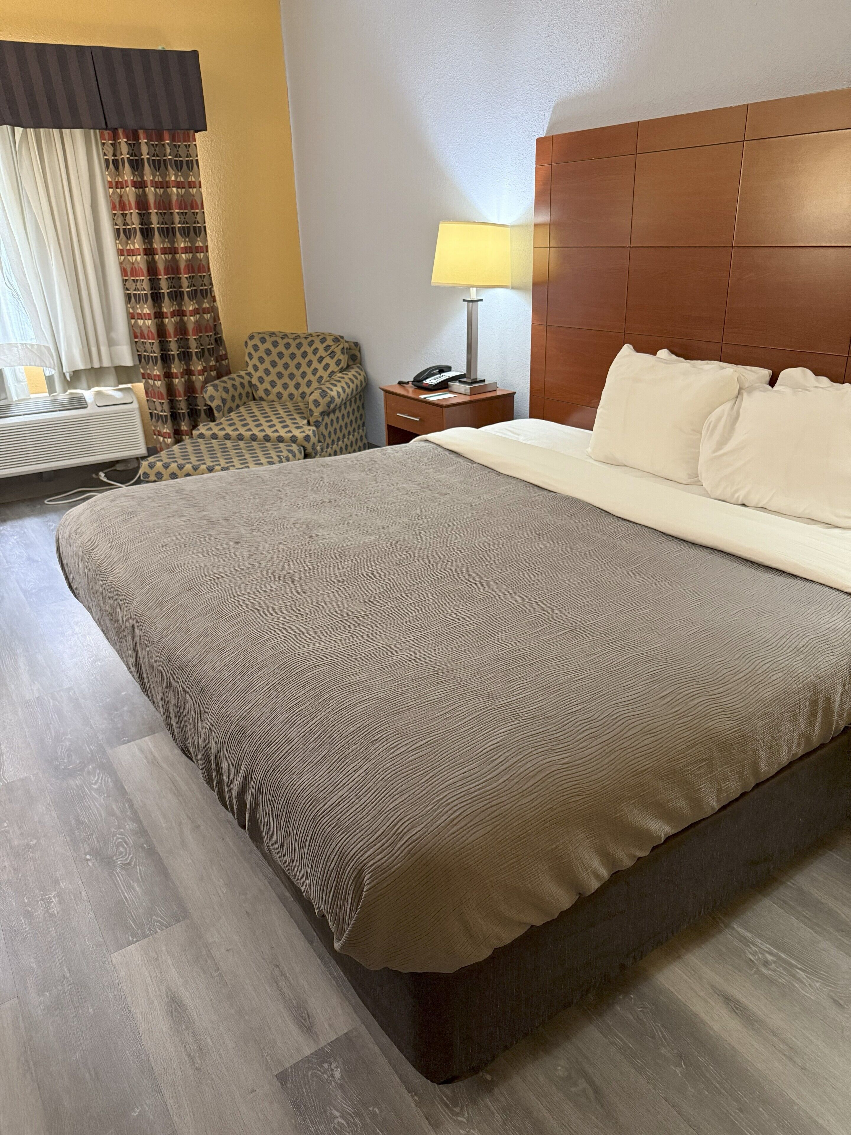Una habitación de hotel con una cama grande, una mesita de noche con lámpara, una silla y una ventana con cortinas.