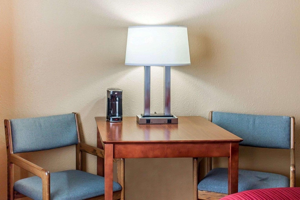 Quarto de hotel com uma mesa de madeira e duas cadeiras azuis com almofadas. Uma luminária de mesa e uma cafeteira estão sobre a mesa.