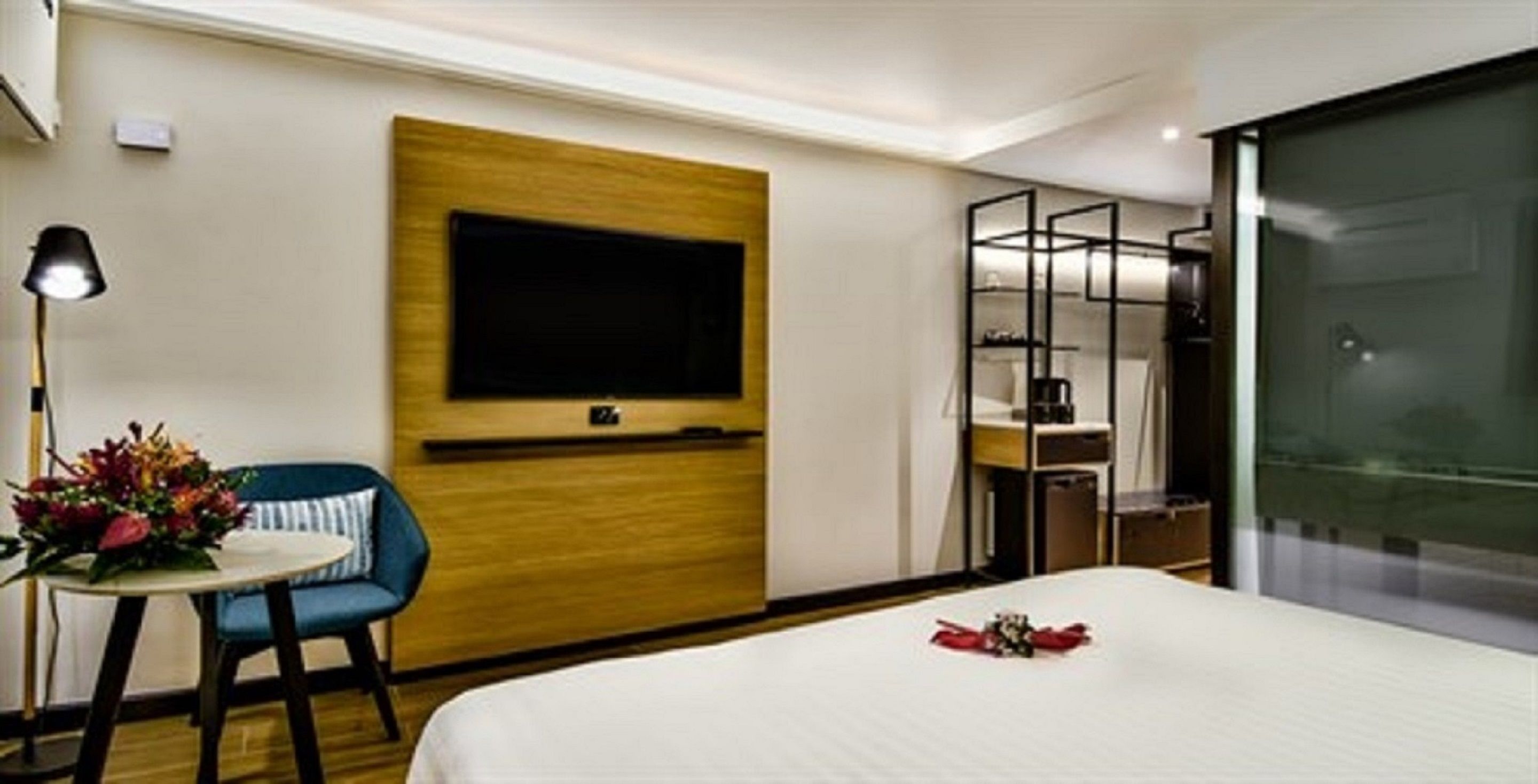 A hotel room with a bed, a desk with a chair, a TV mounted on the wall, and a glass partition.